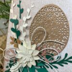 wielkanocna kartka okolicznościowa z brązową pisanką ozdobiona zielonymi listkami i białym kwiatkiem