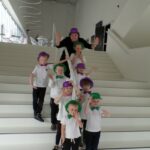 ośmioro dzieci z nauczycielką stoi na białych schodach wszyscy mają ubrane kolorowe muszki i na głowach kolorowe kapelusze
