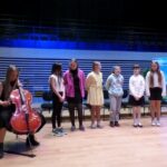 uczennica przygotowuje wiolonczelę do gry siedząc na scenie za nią stoi w rzędzie sześć dziewczynek
