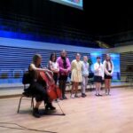 uczennica gra na wiolonczeli siedząc na scenie za nią stoi w rzędzie sześć dziewczynek jedna z nich trzyma w ręce mikrofon