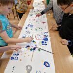 dzieci stojąc przy długim stole malują farbami kolorowe kropki
