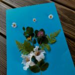 płaska praca plastyczna wykonana na niebieskiej kartce przyklejone są naturalne kwiatki i liście