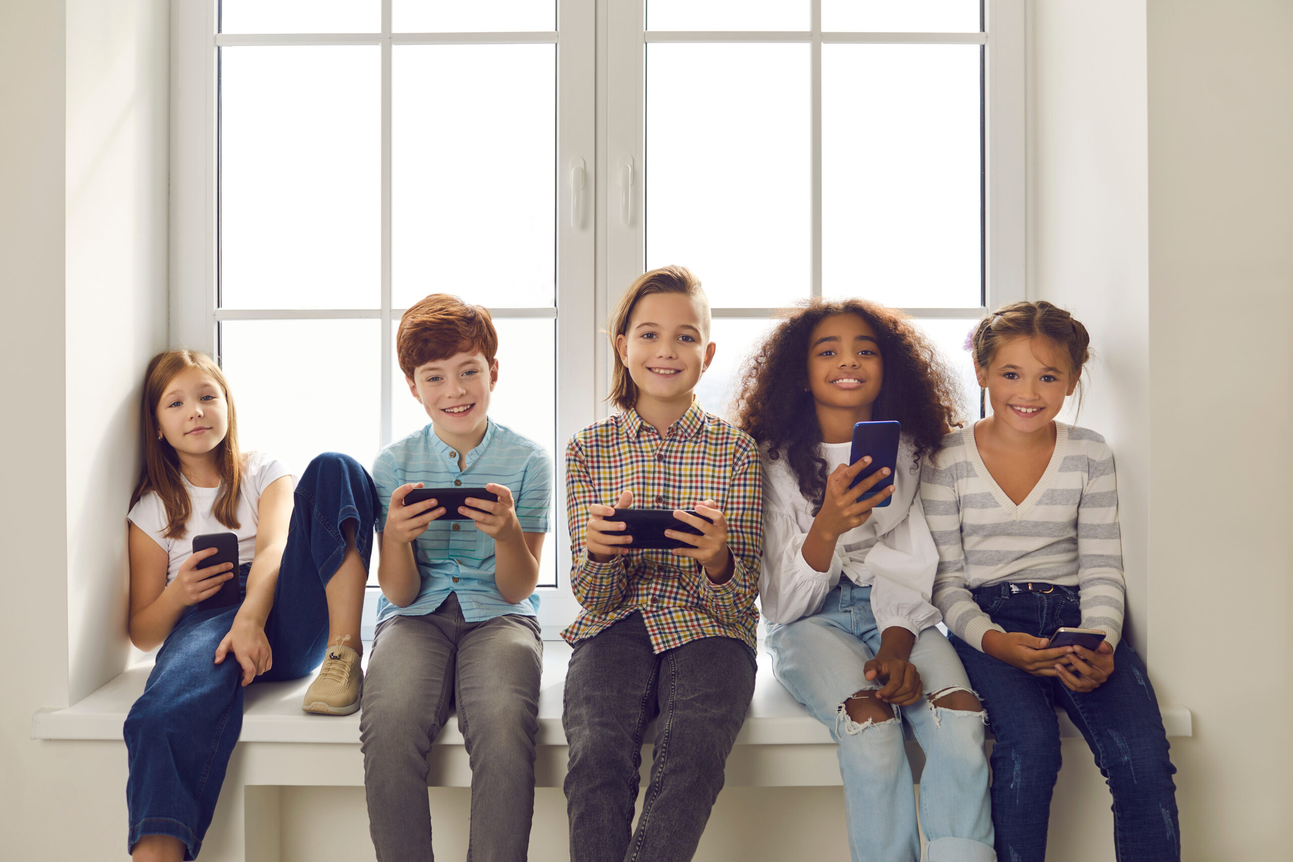 Diverse group of happy 9-10 year old children playing online games on cell phones. Smiling boys and girls holding mobile devices and looking at camera sitting on windowsill with white mockup window uśmiechnięte dzieci z telefonami w dłoniach siedzą na parapecie