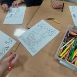 zdjęcie przedstawia stolik na którym są kolorowe kredki w pudełku oraz ręce uczniów wykonujących tematyczne prace plastyczne