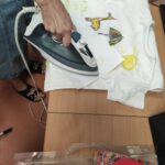 Zdjęcie przedstawia rękę nauczyciela prasującego koszulkę przez kartkę w celu utrwalenia namalowanego przez ucznia na bluzce wzoru.