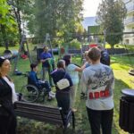uczniowie wraz z nauczycielami stoją na placu zabaw i słuchają instrukcji oraz odbierają od nauczycielki rękawiczki jednorazowe