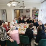 Uczniowie wraz z nauczycielem i pensjonariuszami domu pomocy społecznej wspólnie siedzą przy stole.