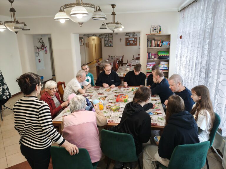 Uczniowie wraz z nauczycielem i pensjonariuszami domu pomocy społecznej wspólnie siedzą przy stole.